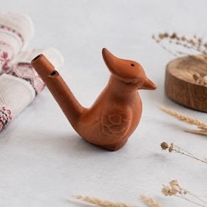 May include: A terracotta-colored clay bird whistle featuring a stylized avian shape with a pointed beak, a small hole on the head, and a tubular mouthpiece extending from the back. The surface displays a matte finish with carved wing details.