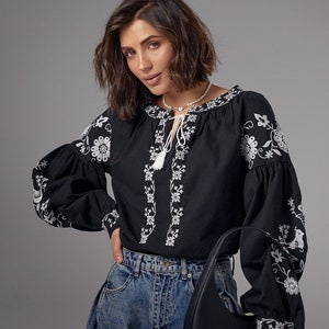 May include: A black embroidered blouse with white floral designs and a white tassel tie. The blouse has long sleeves and a relaxed fit. The model is wearing blue jeans and a black handbag.
