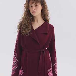 May include: A deep burgundy wrap cardigan featuring a wide collar and a matching tie belt at the waist. The long sleeves and lower sides of the garment are decorated with a geometric star pattern in shades of light pink and white.