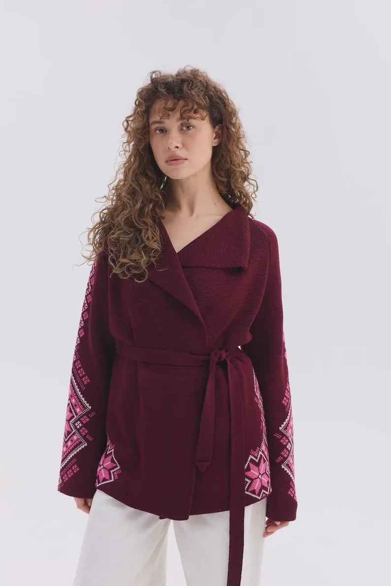 May include: A deep burgundy wrap cardigan featuring a wide collar and a matching tie belt at the waist. The long sleeves and lower sides of the garment are decorated with a geometric star pattern in shades of light pink and white.