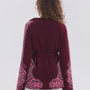 May include: A deep burgundy knit cardigan featuring long sleeves and a waist tie. The garment is decorated with a repeating geometric star pattern in shades of light pink and white along the sleeves and the lower back hemline.