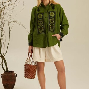 Puede incluir: Una camisa de manga larga color verde oliva con paneles rectangulares negros y bordados florales, combinada con pantalones cortos color crema, sandalias negras y un bolso tipo cubo trenzado marr&oacute;n con red.