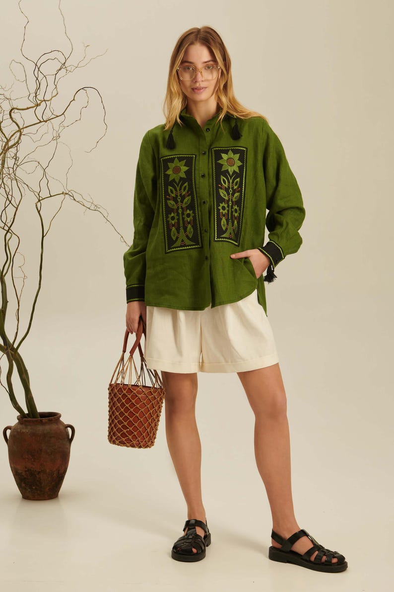 Puede incluir: Una camisa de manga larga color verde oliva con paneles rectangulares negros y bordados florales, combinada con pantalones cortos color crema, sandalias negras y un bolso tipo cubo trenzado marr&oacute;n con red.
