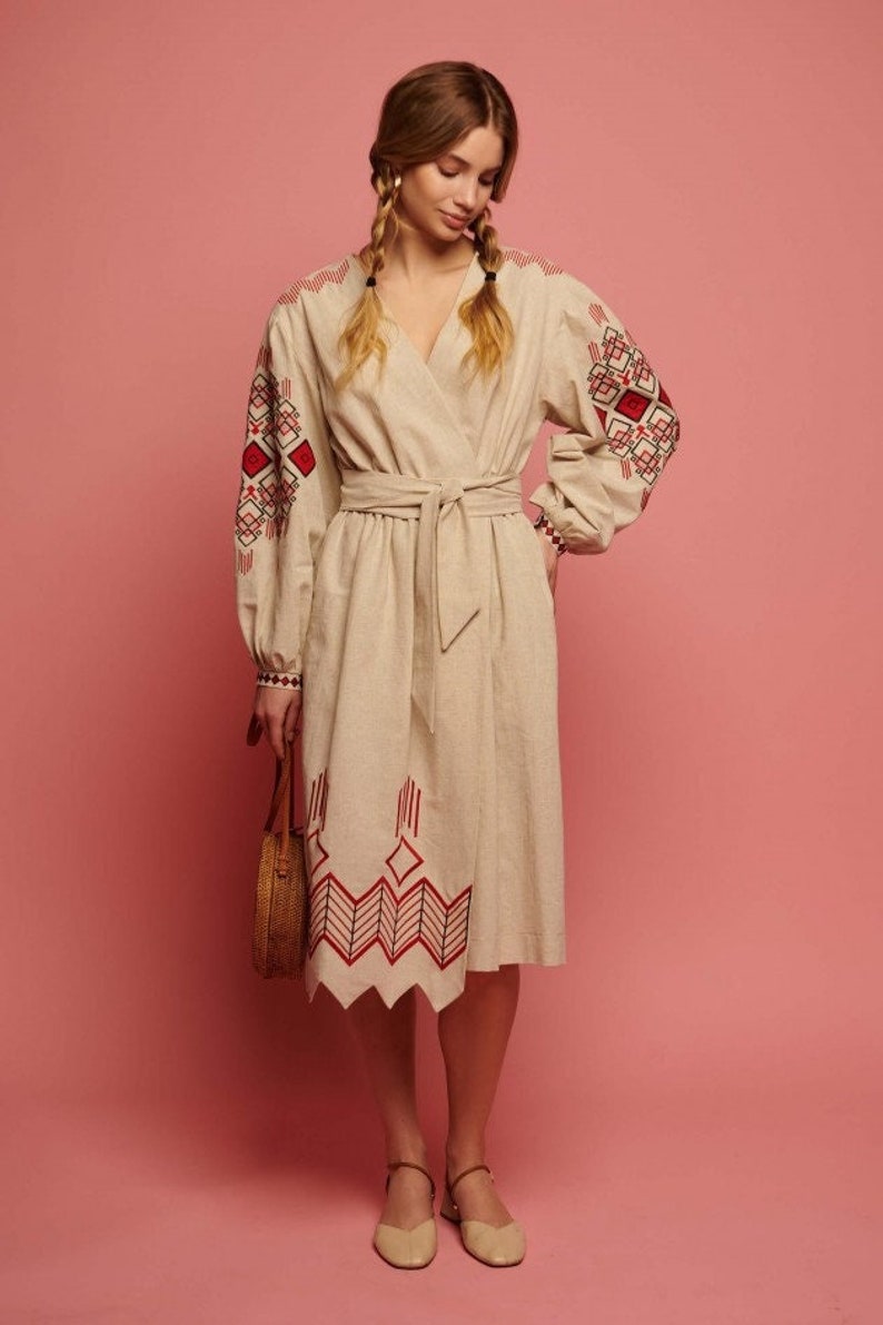 May include: A beige linen dress with red and black embroidered geometric patterns on the sleeves and hem. The dress has a wrap style and a belt.