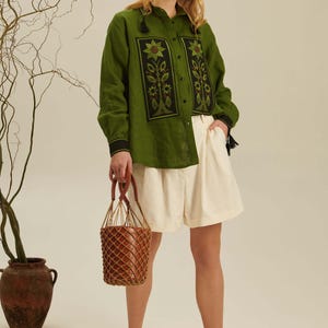 Puede incluir: Una camisa de manga larga color verde oliva con paneles rectangulares negros y motivos de girasoles bordados, combinada con pantalones cortos plisados color crema, sandalias negras de tiras y un bolso tipo cubo marr&oacute;n con red.