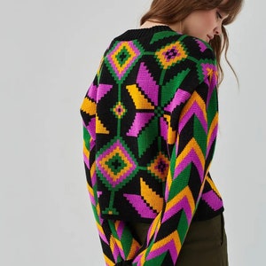 Puede incluir: Un su&eacute;ter negro con un patr&oacute;n geom&eacute;trico en tonos de rosa, verde y amarillo. El su&eacute;ter tiene cuello redondo y mangas largas.