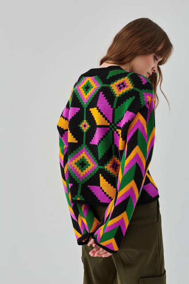 Puede incluir: Un su&eacute;ter negro con un patr&oacute;n geom&eacute;trico en tonos de rosa, verde y amarillo. El su&eacute;ter tiene cuello redondo y mangas largas.