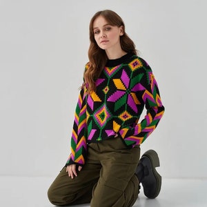 Puede incluir: Una mujer lleva un su&eacute;ter negro con un patr&oacute;n geom&eacute;trico colorido. El su&eacute;ter tiene detalles en rosa, amarillo y verde. Tambi&eacute;n lleva pantalones cargo verde oliva y botas negras.