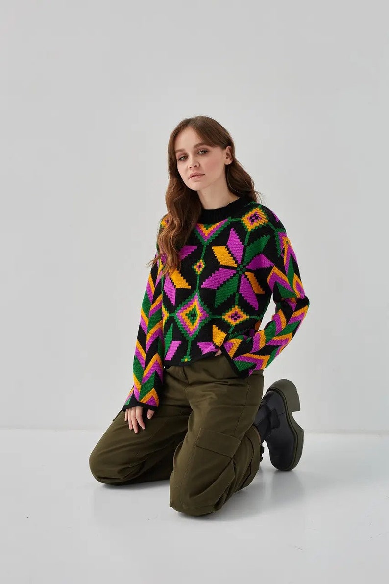 Puede incluir: Una mujer lleva un su&eacute;ter negro con un patr&oacute;n geom&eacute;trico colorido. El su&eacute;ter tiene detalles en rosa, amarillo y verde. Tambi&eacute;n lleva pantalones cargo verde oliva y botas negras.