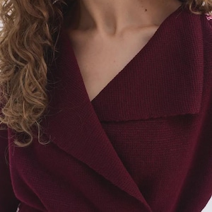 May include: A deep burgundy knit wrap cardigan featuring a wide folded collar, long sleeves, and a matching fabric tie belt at the waist. The garment is crafted from a textured knit material with a visible weave, providing a structured yet soft appearance.