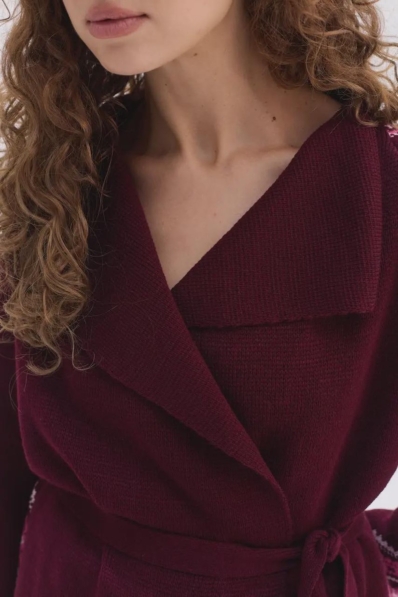 May include: A deep burgundy knit wrap cardigan featuring a wide folded collar, long sleeves, and a matching fabric tie belt at the waist. The garment is crafted from a textured knit material with a visible weave, providing a structured yet soft appearance.