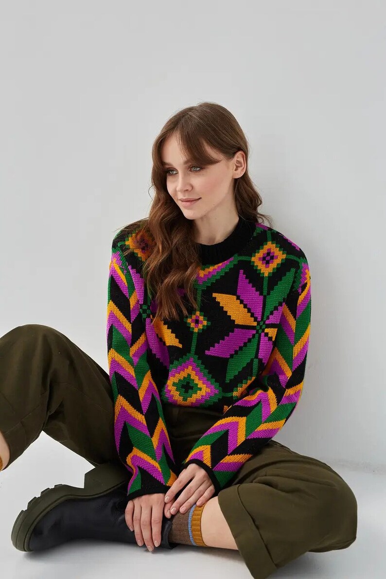 Puede incluir: Un su&eacute;ter negro con un patr&oacute;n geom&eacute;trico colorido en tonos de verde, morado, amarillo y naranja. El su&eacute;ter tiene mangas largas y cuello redondo.