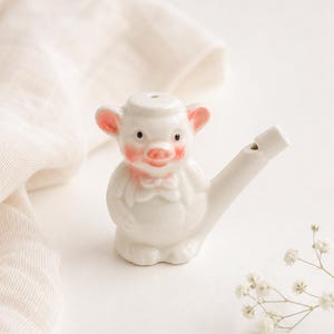 May include: A white ceramic whistle shaped like a pig wearing a small hat and a bow tie. The pig features hand-painted pink accents on its ears, cheeks, and snout, with black eyes. A cylindrical mouthpiece extends from the right side of the figure.