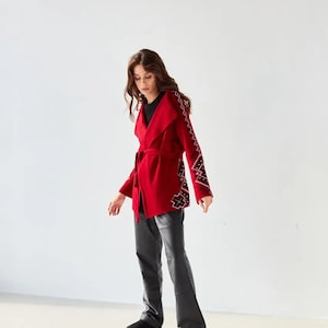 May include: A vibrant red wrap jacket featuring a wide collar, a matching tie belt at the waist, and long sleeves adorned with intricate black and white geometric embroidery patterns along the outer arms, paired with black trousers and black shoes.