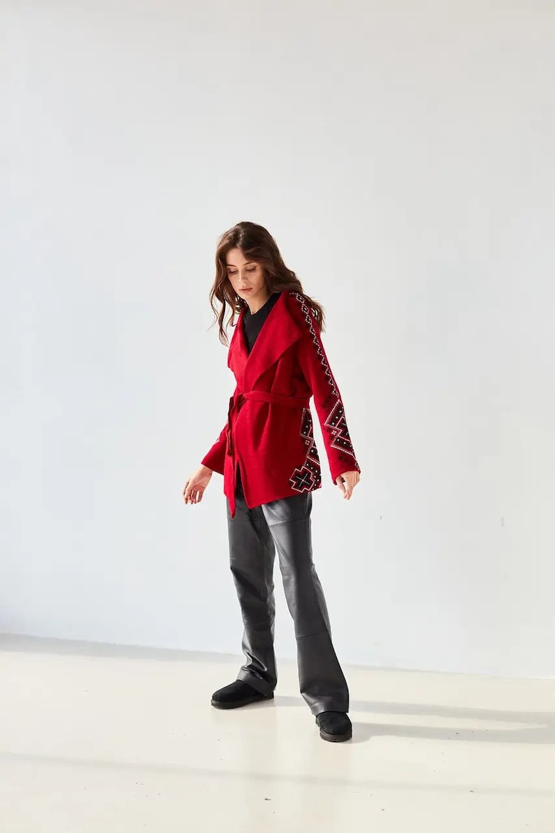May include: A vibrant red wrap jacket featuring a wide collar, a matching tie belt at the waist, and long sleeves adorned with intricate black and white geometric embroidery patterns along the outer arms, paired with black trousers and black shoes.