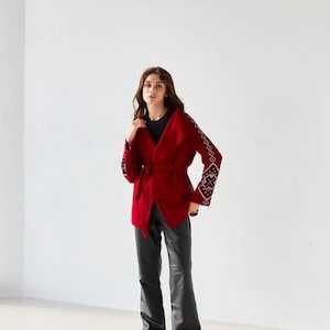 May include: A deep red wrap jacket featuring a wide collar, a matching tie belt at the waist, and long sleeves adorned with intricate black and white geometric embroidery patterns. The jacket is paired with black trousers and black slip-on shoes.