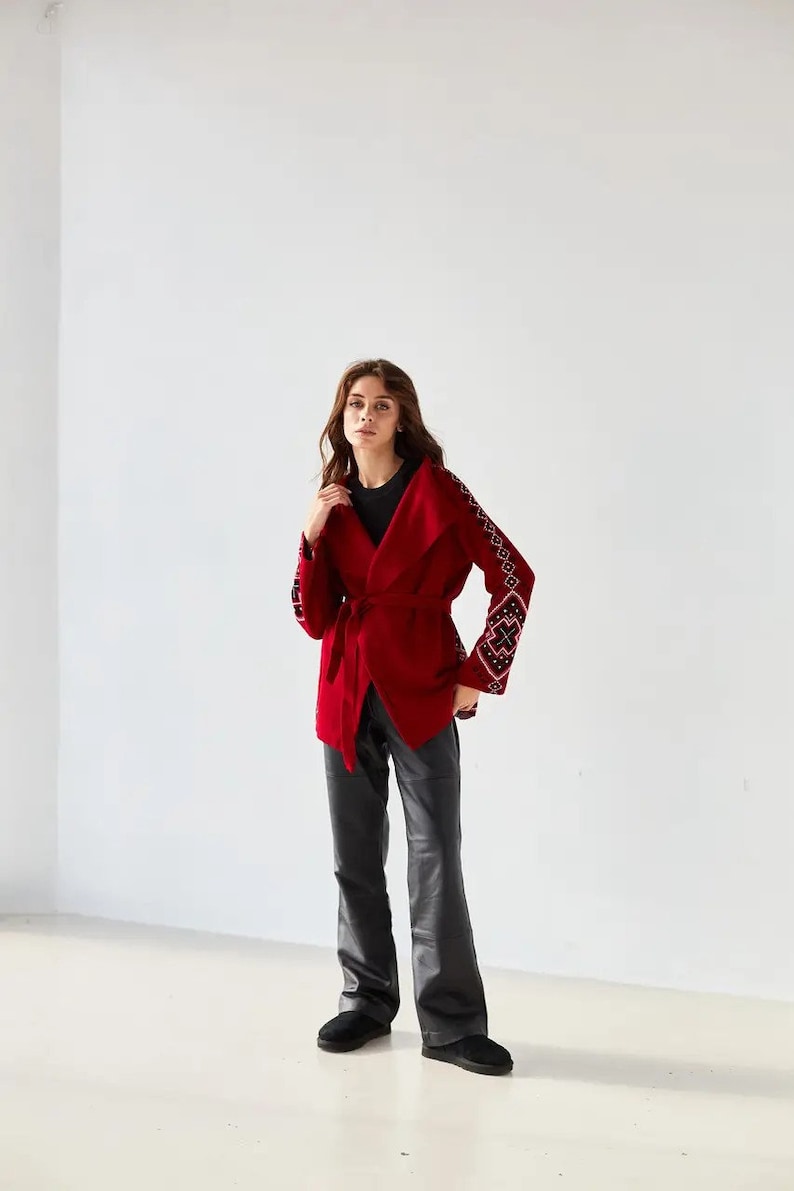 May include: A deep red wrap jacket featuring a wide collar, a matching tie belt at the waist, and long sleeves adorned with intricate black and white geometric embroidery patterns. The jacket is paired with black trousers and black slip-on shoes.