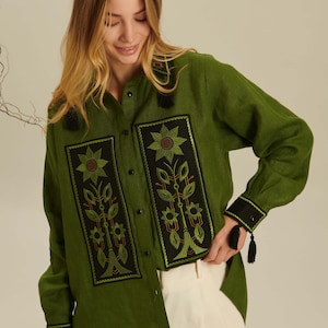 Puede incluir: Una camisa de manga larga color verde oliva con botones y dos paneles rectangulares negros en la parte delantera con bordados florales en verde y naranja. La camisa incluye cuello, botones negros y pu&ntilde;os negros decorados con borlas peque&ntilde;as.