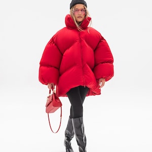 Peut inclure: Une doudoune rouge vif oversize avec un col montant et une fermeture éclair, associée à un legging noir, des bottes noires arrivant au genou, un bonnet gris foncé, des lunettes teintées et un petit sac à main rouge structuré à fermoir doré.