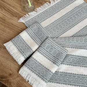 May include: A rectangular table runner and matching placemat featuring a woven pattern of sage green geometric shapes and stripes on an off-white background. Both pieces are finished with short, white fringe along the edges.