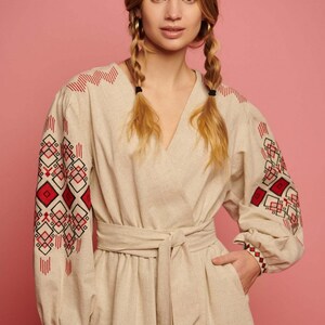 May include: A beige linen dress with red and black embroidered geometric patterns on the sleeves. The dress has a V-neckline and a tie belt at the waist.