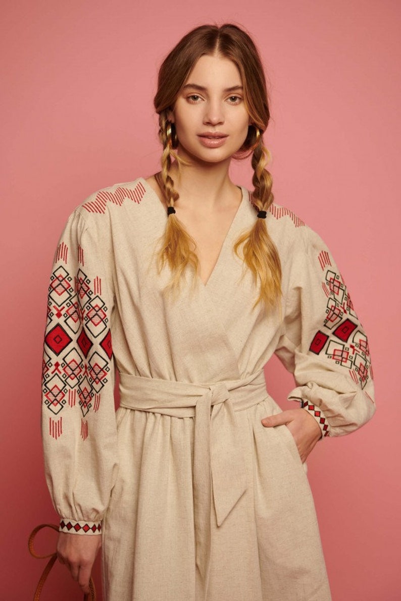 May include: A beige linen dress with red and black embroidered geometric patterns on the sleeves. The dress has a V-neckline and a tie belt at the waist.