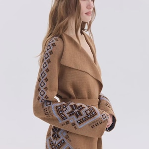 May include: A tan knit cardigan featuring a wide shawl collar, a tie waist, and long sleeves. The garment is decorated with a blue and dark brown geometric snowflake pattern along the sleeves and the lower front panels of the knit fabric.