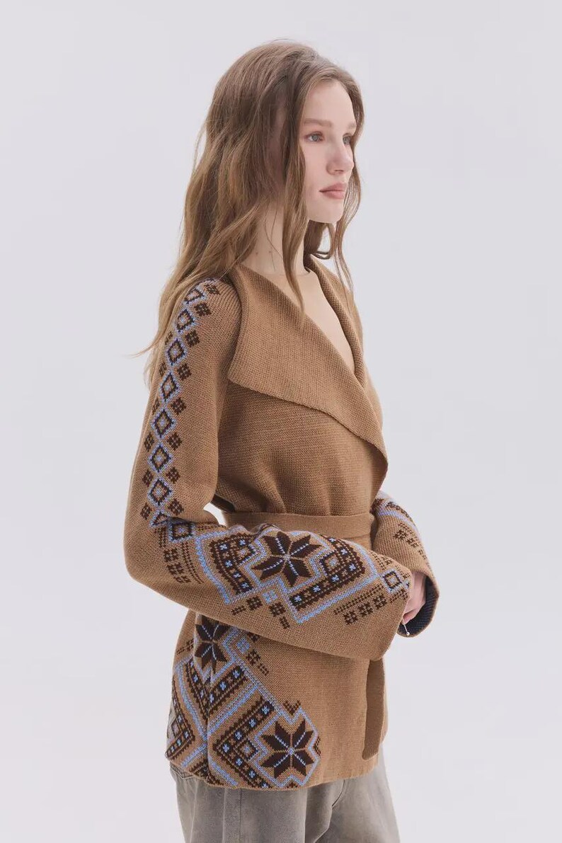 May include: A tan knit cardigan featuring a wide shawl collar, a tie waist, and long sleeves. The garment is decorated with a blue and dark brown geometric snowflake pattern along the sleeves and the lower front panels of the knit fabric.