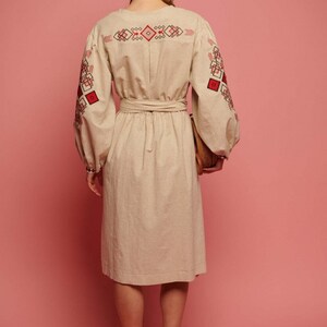 May include: A beige linen dress with red and black embroidered geometric patterns on the sleeves and back. The dress has a cinched waist and a midi length.