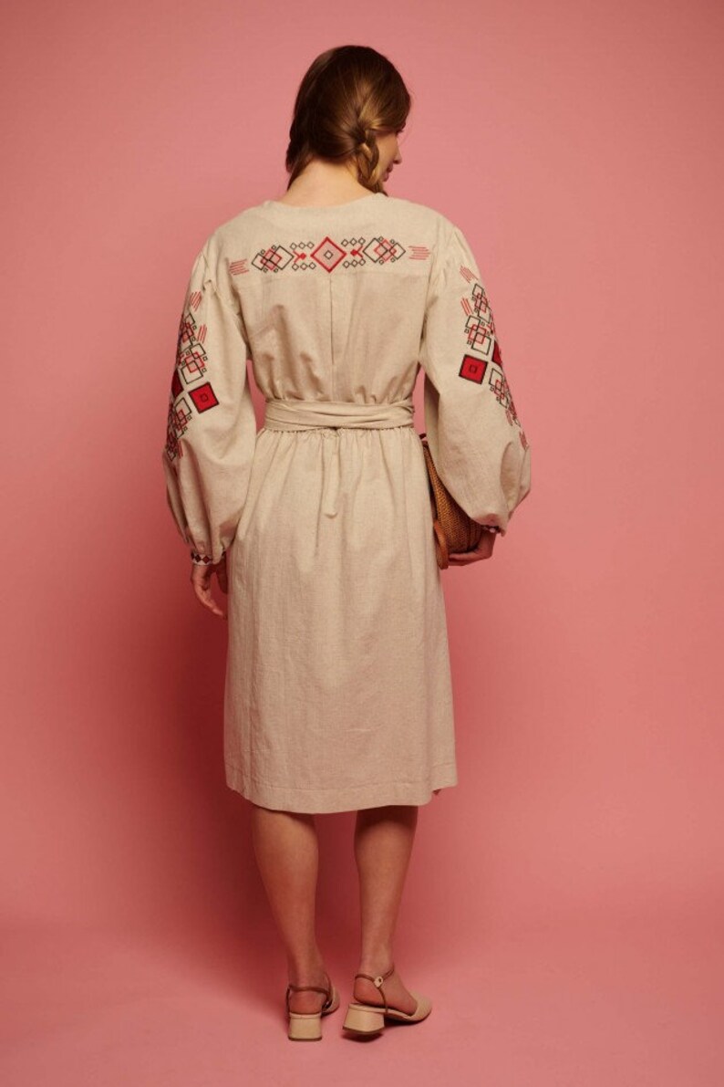 May include: A beige linen dress with red and black embroidered geometric patterns on the sleeves and back. The dress has a cinched waist and a midi length.