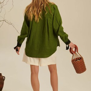 Puede incluir: Una camisa de manga larga verde oliva con espalda plisada, pu&ntilde;os negros con peque&ntilde;as borlas y dobladillo curvo. La camisa se combina con pantalones cortos color crema y sandalias negras, junto a un bolso tipo cubo marr&oacute;n con red.