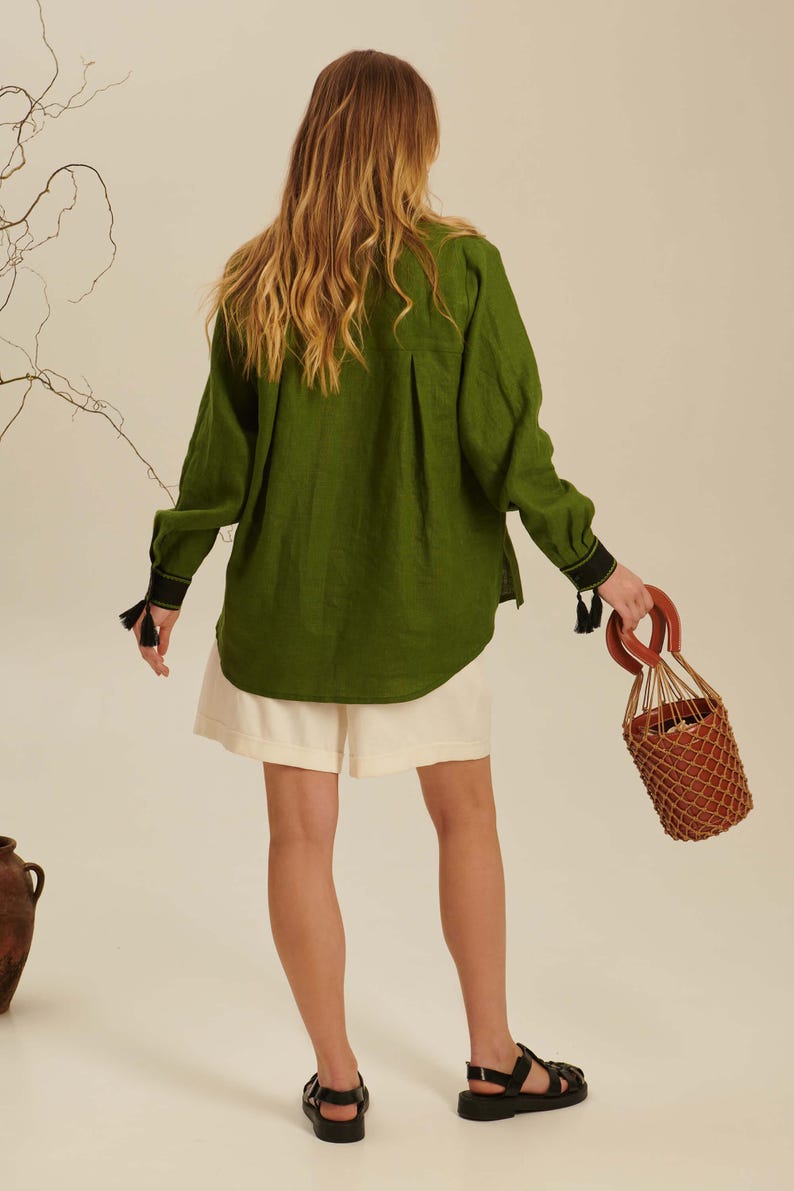 Puede incluir: Una camisa de manga larga verde oliva con espalda plisada, pu&ntilde;os negros con peque&ntilde;as borlas y dobladillo curvo. La camisa se combina con pantalones cortos color crema y sandalias negras, junto a un bolso tipo cubo marr&oacute;n con red.