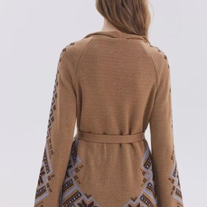 May include: A tan knit cardigan featuring long sleeves and a waist tie. The garment is decorated with a blue, brown, and white geometric snowflake pattern along the lower back and down the length of the outer sleeves, set against a solid tan background.