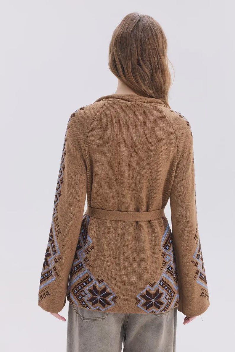 May include: A tan knit cardigan featuring long sleeves and a waist tie. The garment is decorated with a blue, brown, and white geometric snowflake pattern along the lower back and down the length of the outer sleeves, set against a solid tan background.
