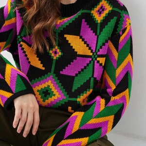 Puede incluir: Un su&eacute;ter negro con un patr&oacute;n geom&eacute;trico colorido en tonos de verde, morado y amarillo. El su&eacute;ter tiene cuello redondo y mangas largas.
