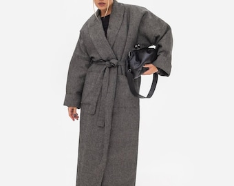 May include: A long, charcoal gray wool-blend coat featuring a wide lapel, long sleeves with folded cuffs, and a matching tie belt at the waist. The coat is paired with a black leather clutch bag, black sunglasses, white socks, and black sandals.