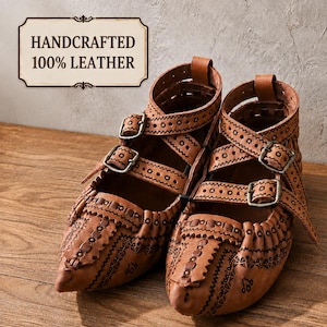 Puede incluir: Un par de zapatos de cuero color canela con intrincados patrones negros estampados, puntas afiladas y dos correas ajustables con hebillas metálicas. Un letrero rectangular arriba muestra el texto HANDCRAFTED 100% LEATHER.
