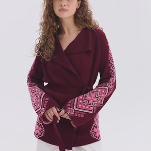 May include: A deep burgundy wrap cardigan featuring a wide collar, a tie waist belt, and long sleeves adorned with intricate geometric star patterns in shades of light pink, white, and dark pink, paired with plain white trousers.
