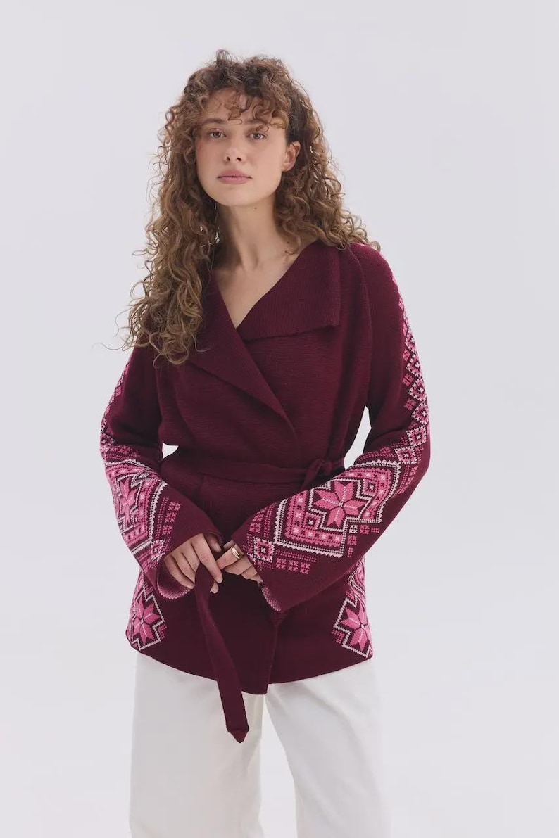 May include: A deep burgundy wrap cardigan featuring a wide collar, a tie waist belt, and long sleeves adorned with intricate geometric star patterns in shades of light pink, white, and dark pink, paired with plain white trousers.