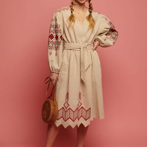 May include: A beige linen dress with red and black embroidered geometric patterns on the sleeves and hem. The dress has a V-neckline and a belt. The model is wearing the dress with a round straw bag and beige flats.