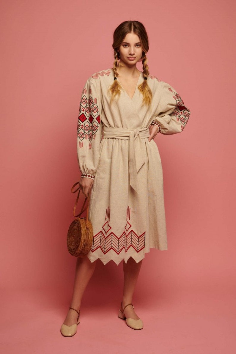 May include: A beige linen dress with red and black embroidered geometric patterns on the sleeves and hem. The dress has a V-neckline and a belt. The model is wearing the dress with a round straw bag and beige flats.