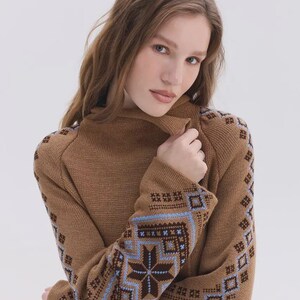 May include: A brown knit sweater featuring a high collar and long sleeves. The sleeves are decorated with a detailed geometric pattern in shades of dark brown, light blue, and white, showcasing intricate star and square motifs throughout.