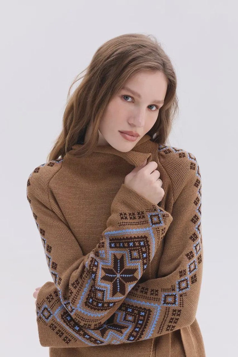 May include: A brown knit sweater featuring a high collar and long sleeves. The sleeves are decorated with a detailed geometric pattern in shades of dark brown, light blue, and white, showcasing intricate star and square motifs throughout.