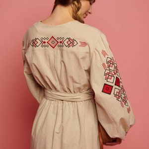 May include: A beige linen dress with red and black embroidered geometric patterns on the sleeves and back. The dress has a cinched waist with a matching fabric belt.