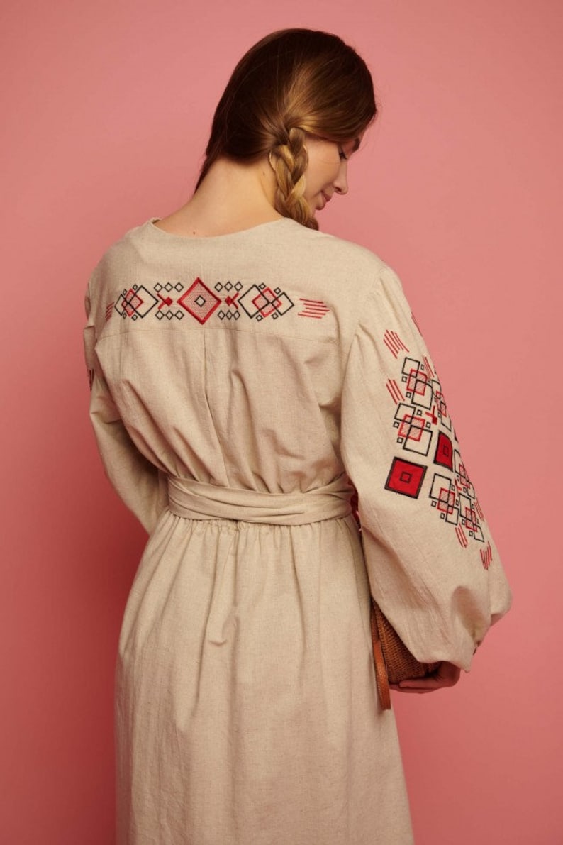 May include: A beige linen dress with red and black embroidered geometric patterns on the sleeves and back. The dress has a cinched waist with a matching fabric belt.