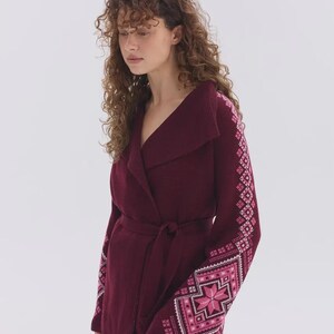 May include: A deep burgundy wrap cardigan featuring a wide collar and a tie waist. The long sleeves are decorated with intricate geometric patterns in shades of pink and white, creating a distinct contrast against the solid dark fabric.