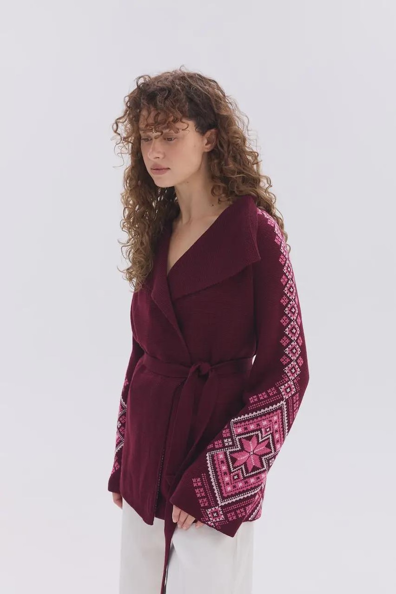 May include: A deep burgundy wrap cardigan featuring a wide collar and a tie waist. The long sleeves are decorated with intricate geometric patterns in shades of pink and white, creating a distinct contrast against the solid dark fabric.