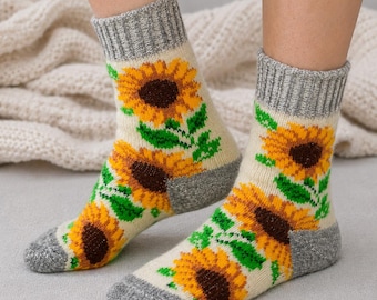 Puede incluir: Un par de calcetines de punto color crema con un patrón repetitivo de girasoles con pétalos amarillos, centros marrón oscuro y hojas verdes. Los calcetines tienen un puño gris acanalado y una sección de talón y puntera gris sólido.