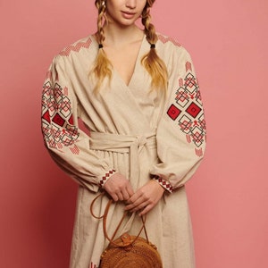 May include: A woman wearing a beige linen dress with red and black embroidered geometric patterns. She is holding a round, brown woven bag with a leather strap.