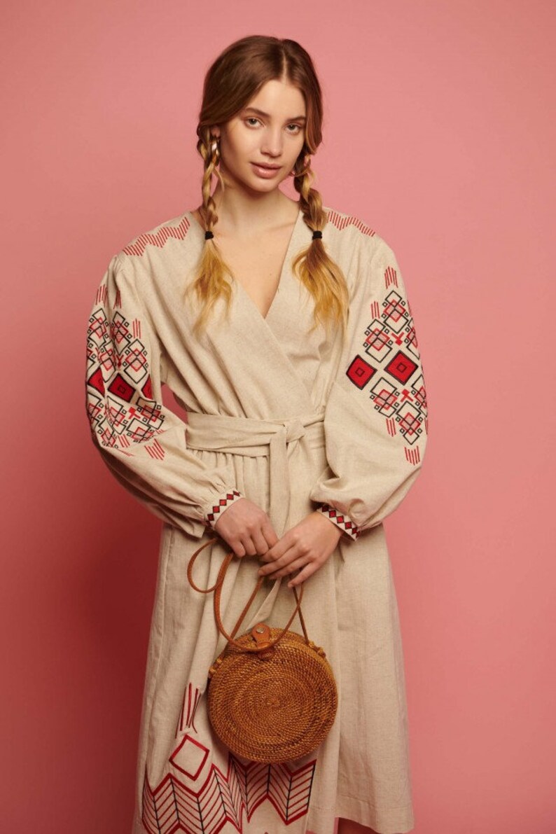 May include: A woman wearing a beige linen dress with red and black embroidered geometric patterns. She is holding a round, brown woven bag with a leather strap.